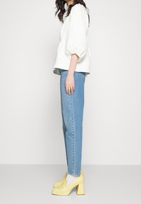 Vit texturerad blus med puffärmar, ljusblå högmidjade jeans och gula blockklackar i konstläder, sedd från sidan.