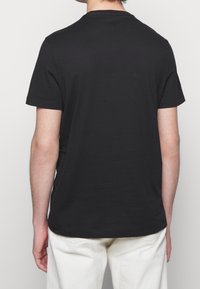 Hombre vestido con una camiseta negra de manga corta sencilla y pantalones blancos, visto desde la espalda contra un fondo claro y liso.