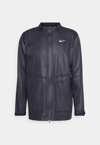 Sort Nike jakke med ståkrave, lynlåslukning og tekstureret stof med diskrete paneldetaljer. Indeholder et lille hvidt logo på brystet.