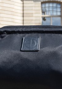 Musta kangaskassi, jossa on ommeltu neliön muotoinen logopatch, jossa on kohokuvioitu pyöreä kuvio, sumennetun ikkunallisen rakennuksen edessä.