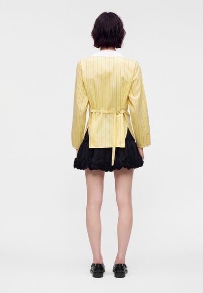 Femme aux cheveux courts et foncés portant un blazer jaune à fines rayures avec ceinture et une mini-jupe noire à volants, debout de dos sur un fond uni.
