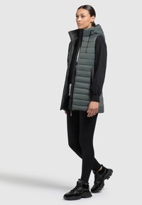 Vadderad väst i mörkgrön med hög krage och öppen framkant, lager på lager över en svart långärmad topp och svarta leggings. Svarta sneakers fulländar looken.