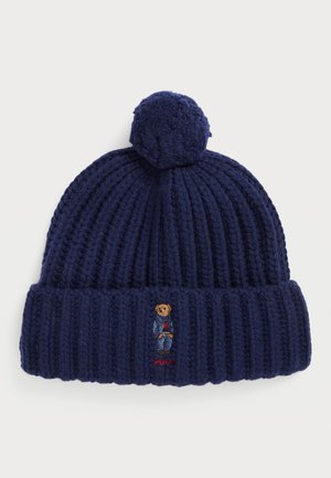 Tumsā zila adīta cepure ar ribotu faktūru, kas vainagota ar pom-pom. Iekļauta izšūta lāča dizains un "Polo" logo.