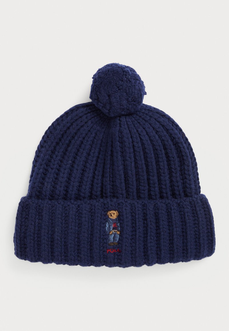 Tumsā zila adīta cepure ar ribotu faktūru, kas vainagota ar pom-pom. Iekļauta izšūta lāča dizains un "Polo" logo.