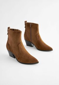 Stivaletti in suede marrone con punta affusolata e tacco basso a blocco. Presentano una cerniera laterale e un'etichetta in tessuto sul retro.