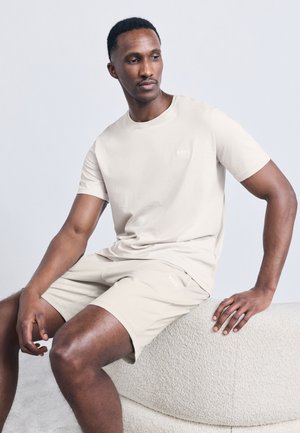 Άνδρας που φοράει μπεζ κοντομάνικο πουκάμισο και σορτς ταιριαστό, καθισμένος σε καμπύλο πάγκο με υφή, μπροστά σε απλό γκρι φόντο.