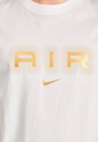 Bijela pamučna majica kratkih rukava s istaknutim zlatnim natpisom "AIR" i malim zlatnim Nike swoosh logom. Jednostavna okrugla izreza i kratki rukavi.