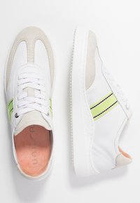 Baskets blanches avec accents en daim, présentant des détails à bandes vert néon et noir, semelle en caoutchouc texturée, et lacets blancs.