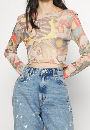 Femme portant un crop top transparent à manches longues avec des imprimés pop art colorés et un jean taille haute bleu éclaboussé de peinture blanche.