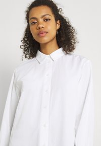 Camicia bianca con bottoni realizzata in tessuto leggero, dotata di colletto a punta, maniche lunghe e chiusura con bottoni frontali. Texture liscia.