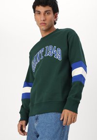 Groene sweatshirt met tekst "GANT 1949", voorzien van blauwe en crèmekleurige gestreepte accenten op de mouwen. Gemaakt van zachte stof met een relaxed fit.