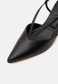 Chaussure plate noire à bout pointu avec fines brides croisées à la cheville et dessus en cuir synthétique texturé.