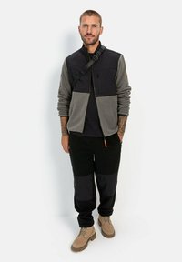 Fleecejacke mit grauen und schwarzen Farbbereichen, mit Reißverschluss-Brusttasche, kombiniert mit schwarzen Hosen und braunen Stiefeln. Mit Schultergurt.