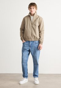 Lys beige lynlåste jakke med høj krave, elastisk talje og manchetter, kombineret med lyseblå jeans og hvide sneakers.