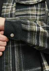 Cămașă din flanel cu carouri verzi și negre, cu un finisaj texturat, având un buton metalic argintiu pe manșetă și detalii albe în contrast.
