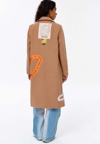 Žena v kabátu z velbloudí srsti s grafickými nášivkami a textem, v kombinaci se světle modrými širokými džíny a bílými teniskami, stojící zády.