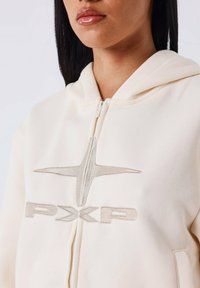 Felpa color crema con zip completa, caratterizzata da un logo e una grafica ricamati in argento sul davanti. Tessuto morbido e cappuccio regolabile.