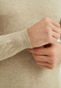 Poignet d'un pull en tricot beige mettant en valeur une texture côtelée. Mains ajustant la manche, démontrant un tissu léger et doux.