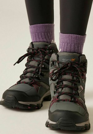 Botas de senderismo grises y negras con cordones con acentos rojos, usadas con calcetines de punto morados y pantalones negros sobre un fondo neutro.