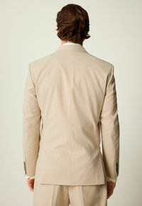 Chaqueta de traje beis con un corte ajustado, abertura en la parte trasera y puños de cuatro botones. Textura suave y solapas para un aspecto formal.