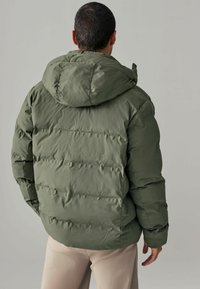 Jachetă puffer verde cu glugă, având modele cu matlasare orizontală și o textură netedă. Modelul este văzut din spate, cu brațele relaxate.