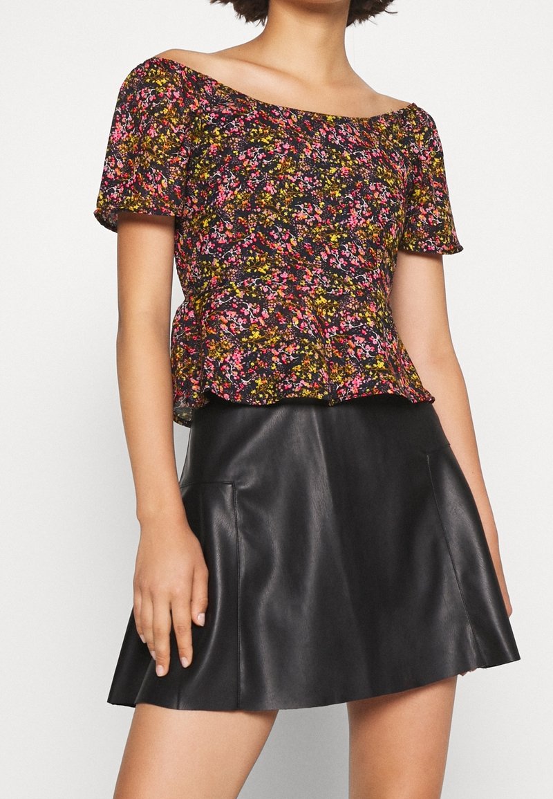 Femme portant un chemisier fleuri à manches courtes et épaules dénudées associé à une jupe en cuir noire de style trapèze.