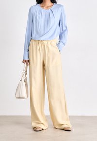 Světlá modrá halenka s dlouhými rukávy a předními záhyby, kombinovaná s volnými, světle žlutými kalhotami a malou bílou kabelkou. V ceně jsou béžové baleríny.