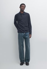 Jeune homme portant un polo bleu marine à manches longues, un jean bleu ample et des chaussures noires, debout devant un fond clair uni.