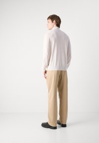 Veste zippée crème à manches longues et légère avec un ourlet côtelé, associée à un pantalon beige et des chaussures noires. Texture lisse, design minimal.