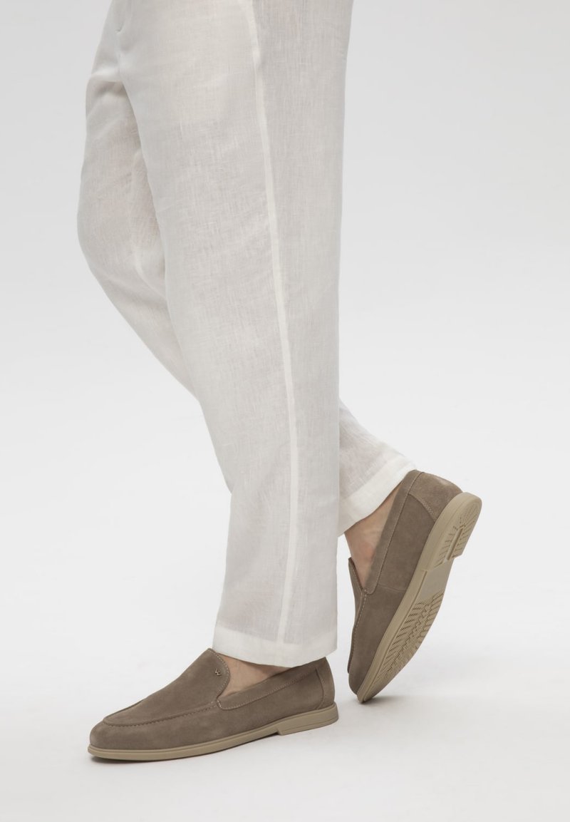 Personne portant un pantalon en lin beige clair et des chaussures en daim marron sans lacets, debout avec un pied légèrement levé.