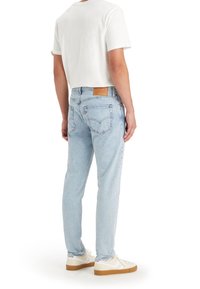 Man i ljusblå jeans, vit t-shirt och vita sneakers står vänd bort från kameran mot en vit bakgrund.