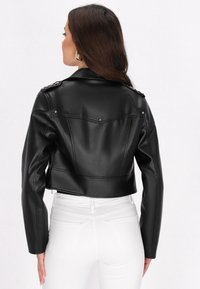 Giubbotto nero cropped in ecopelle con spalle strutturate, borchie metalliche e cuciture sartoriali. Caratterizzato da una superficie liscia e un design elegante.