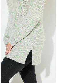 Jasnoszary sweter z dzianiny z zielonymi kropkami, z bocznymi rozcięciami oraz ściągaczami przy mankietach i dolnej krawędzi. Stylizowany z dopasowanymi czarnymi spodniami.