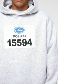 Lichtgrijze hoodie met een grote witte patch met de tekst "POLIZEI 15594" en een blauw logo. De stof lijkt zacht en heeft een standaard pasvorm.