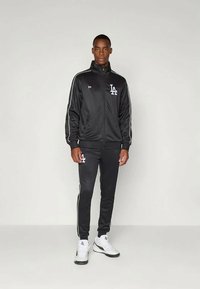 Fekete sportos tréningruha fehér csíkokkal és logóval, cipzáras dzseki, testhezálló nadrág, sima anyagból készült, fehér sportcipővel párosítva.