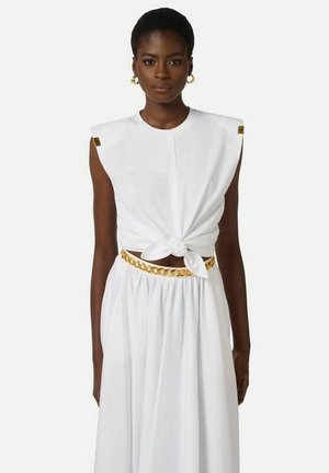 Donna con top bianco senza maniche annodato in vita, gonna bianca con cintura a catena dorata e orecchini a cerchio dorati su sfondo semplice.