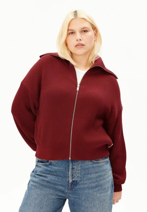 Jonge vrouw met blond haar draagt een geribde bordeauxrode trui met rits over een wit topje en blauwe spijkerbroek, staand tegen een effen achtergrond.