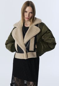 Frau in einer grünen Bomberjacke mit beigem Schaffellkragen und schwarzem Spitzenrock, die vor einem schlichten hellen Hintergrund steht.