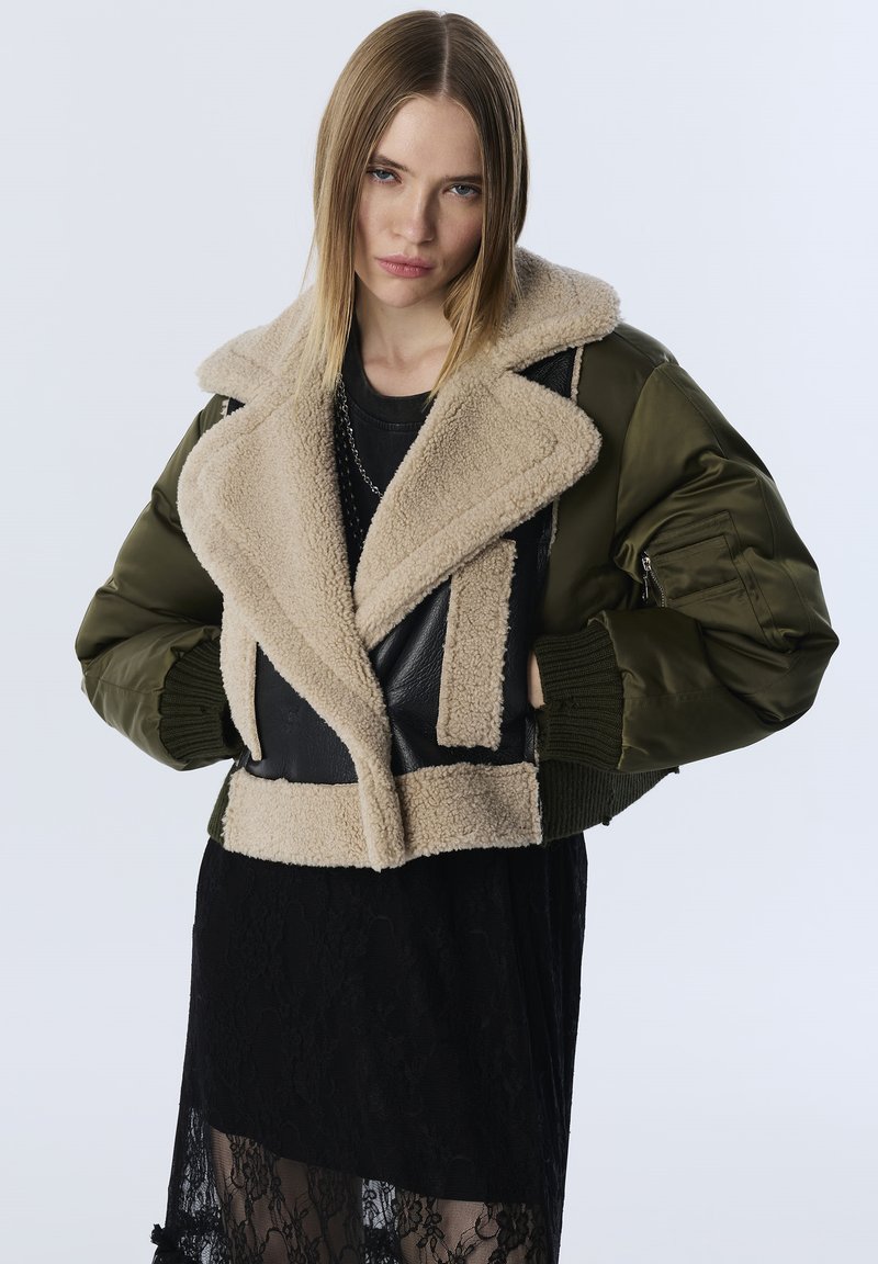 Frau in einer grünen Bomberjacke mit beigem Schaffellkragen und schwarzem Spitzenrock, die vor einem schlichten hellen Hintergrund steht.