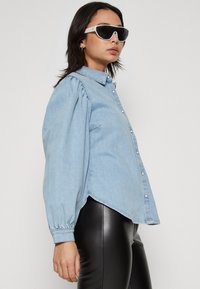 Lys blå denimskjorte med knapper foran, pufferm, og buet hem, kombinert med sorte tettsittende bukser og hvite solbriller.