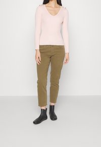 Donna che indossa un maglione a maniche lunghe rosa pallido con scollatura a V, pantaloni verde oliva e stivali neri alla caviglia, in piedi contro uno sfondo chiaro e uniforme.
