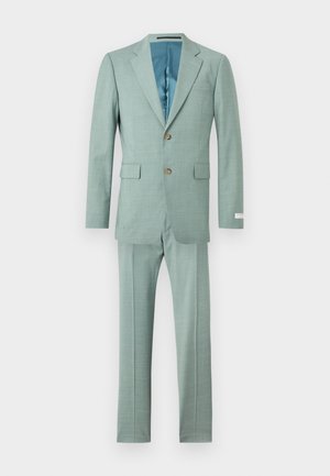 Homme en costume vert menthe avec chemise blanche et cravate noire, debout avec une main dans la poche sur un fond uni.