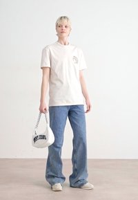 Grietos medvilnės marškinėliai su logotipu, mėlyni plataus kirpimo džinsai, balta rankinė su juodu tekstu ir smėlio spalvos batai. Paprastas fonas.