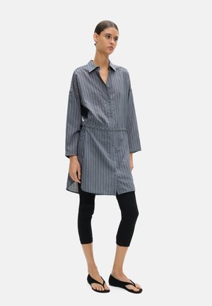 Kobieta stojąca, ubrana w luźną, długorękawową szarą sukienkę koszulową w cienkie paski, czarne krótkie legginsy i czarne klapki na białym tle.