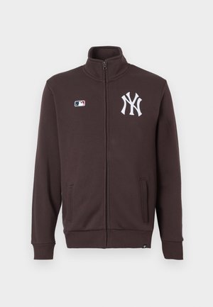 Braune Zip-Jacke aus weichem Material, mit einem hohen Kragen, zwei Vordertaschen und einem weißen Logo auf der linken Brust.