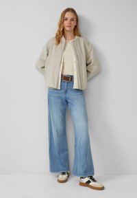 Lichtgrijze wollen bomberjack met ritssluiting, beige top, wijdvallende blauwe jeans en bicolor sneakers met zwarte en crèmekleurige accenten.