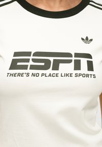 Λευκό αθλητικό πουκάμισο με έντονη μαύρη γραμματοσειρά "ESPN" και τη φράση "ΔΕΝ ΥΠΑΡΧΕΙ ΤΟΠΟΣ ΟΠΩΣ ΤΑ ΑΘΛΗΜΑΤΑ." Ριμπ λαιμός σε πράσινο χρώμα και λογότυπο Adidas.
