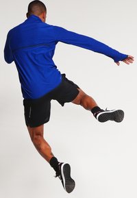 Blå idrottströja med lång ärm och svarta detaljer, tillsammans med svarta shorts och mörka träningsskor, i en dynamisk pose i luften.