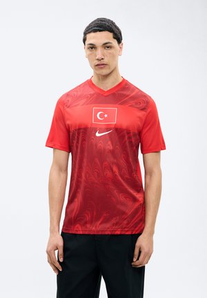 Junger Mann trägt ein rotes Nike-Sporttrikot mit einem wirbelnden Muster und der Flagge der Türkei auf der Brust, steht vor einem einfarbigen Hintergrund.