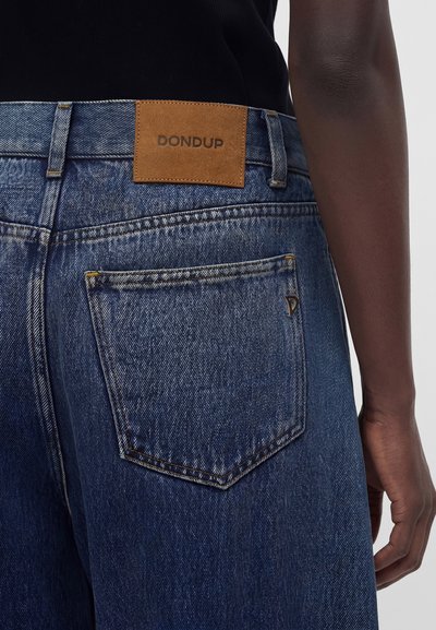 Πίσω όψη ατόμου που φοράει μπλε τζιν denim με καφέ επωνυμία Dondup πάνω από την δεξιά πίσω τσέπη και μαύρο πάνω μέρος.